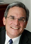 A man with short gray hair and glasses is smiling. He is wearing a dark suit, white shirt, and a patterned tie, and is photographed indoors in front of a blurred background.