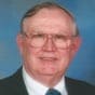 An older man with glasses and short gray hair, wearing a suit and tie, smiles in front of a blue background.