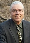 A man with gray slicked-back hair and glasses, wearing a dark suit, olive shirt, and patterned tie, is smiling while sitting outdoors in a wooded area with brown leaves on the ground.