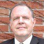 A middle-aged man in a suit and tie is standing in front of a brick wall, looking at the camera with a slight smile.