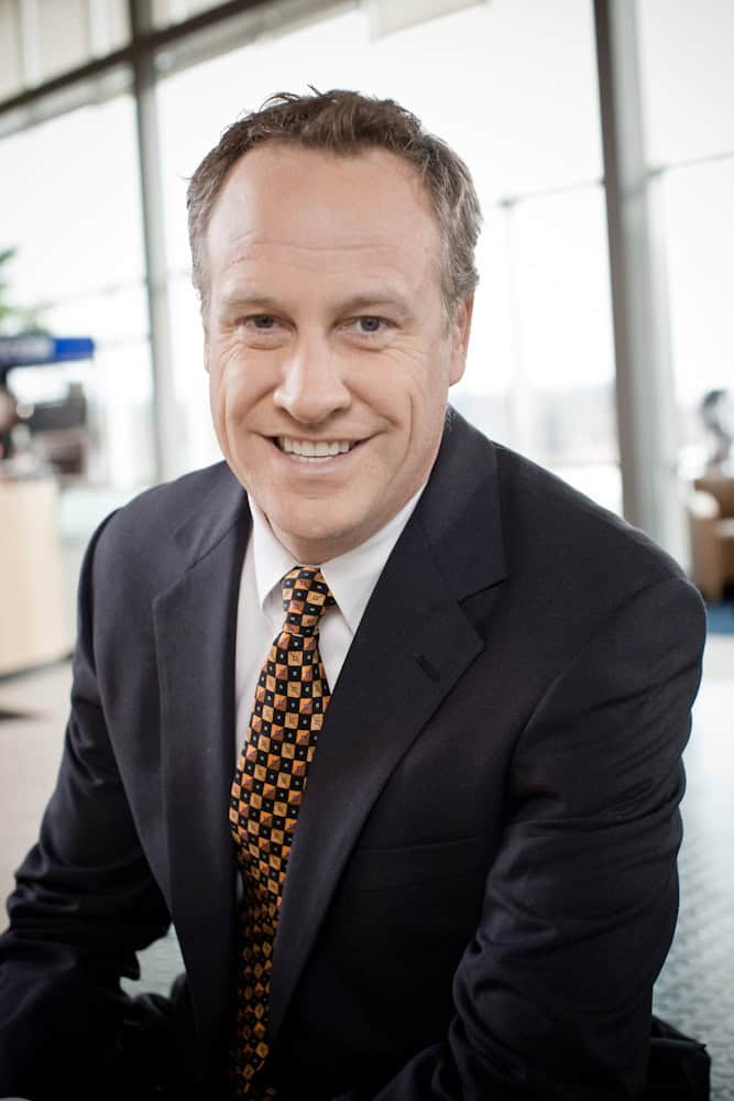 A man in a dark suit and patterned tie smiles at the camera in a modern, bright indoor setting with large windows in the background.