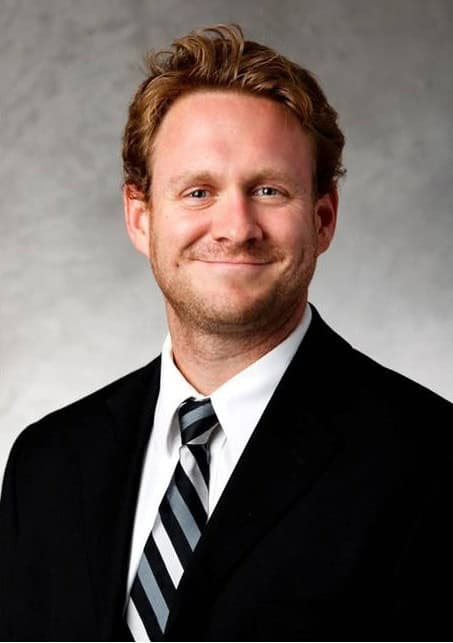A man with short, light brown hair is smiling at the camera. He is wearing a black suit, white dress shirt, and a black-and-white striped tie, with a neutral, light gray background.