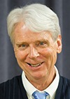 A smiling older man with short, white hair is wearing a black sweater over a white collared shirt and a light blue tie, standing in front of a dark curtain background.