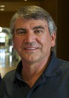 A middle-aged man with gray hair, wearing a dark polo shirt, smiles at the camera indoors, with a blurred background featuring warm lighting and an arched doorway.