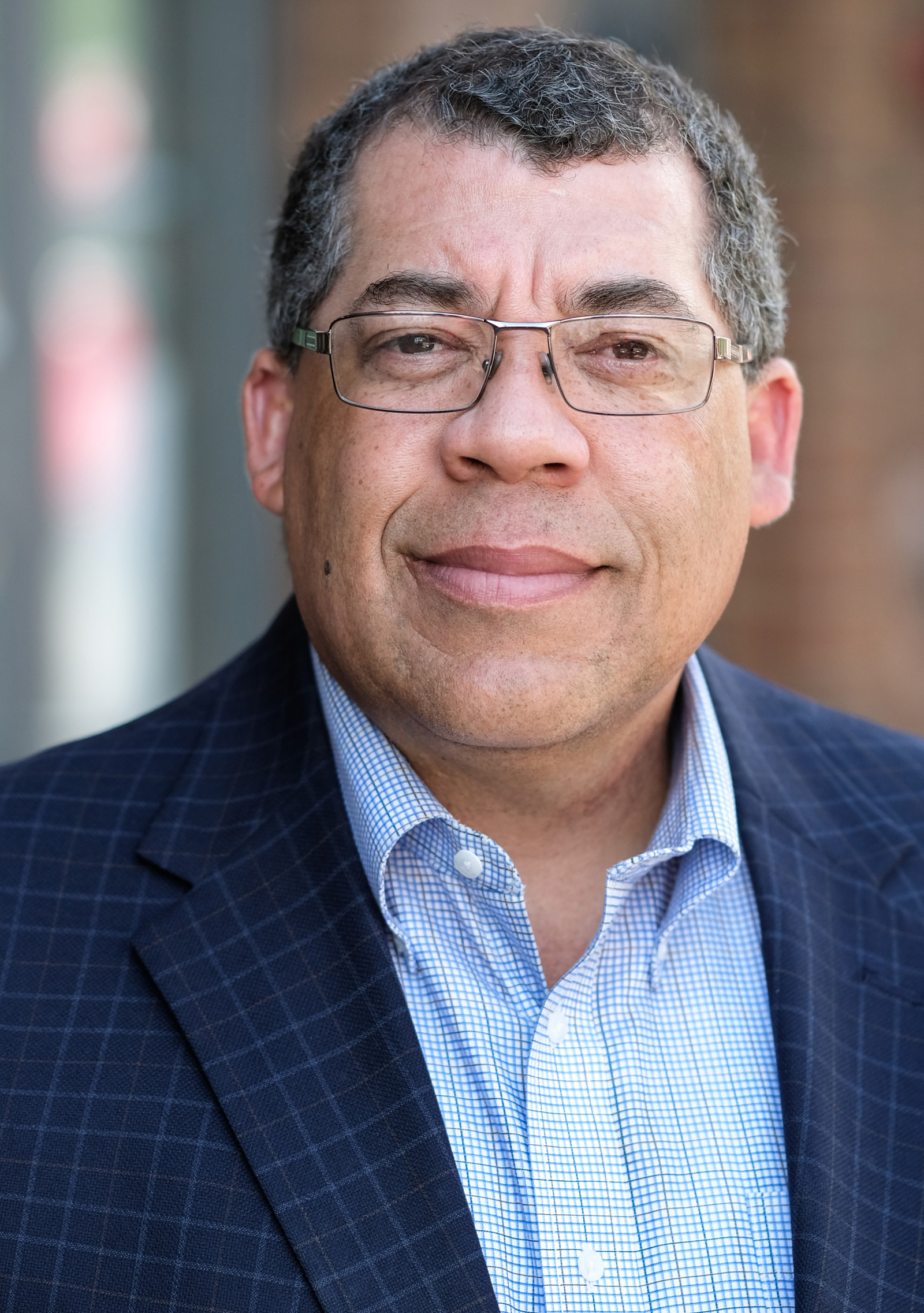 A middle-aged man with short curly hair, glasses, and a slight smile is wearing a blue checkered blazer and a light blue dress shirt, standing outdoors with a blurred background.