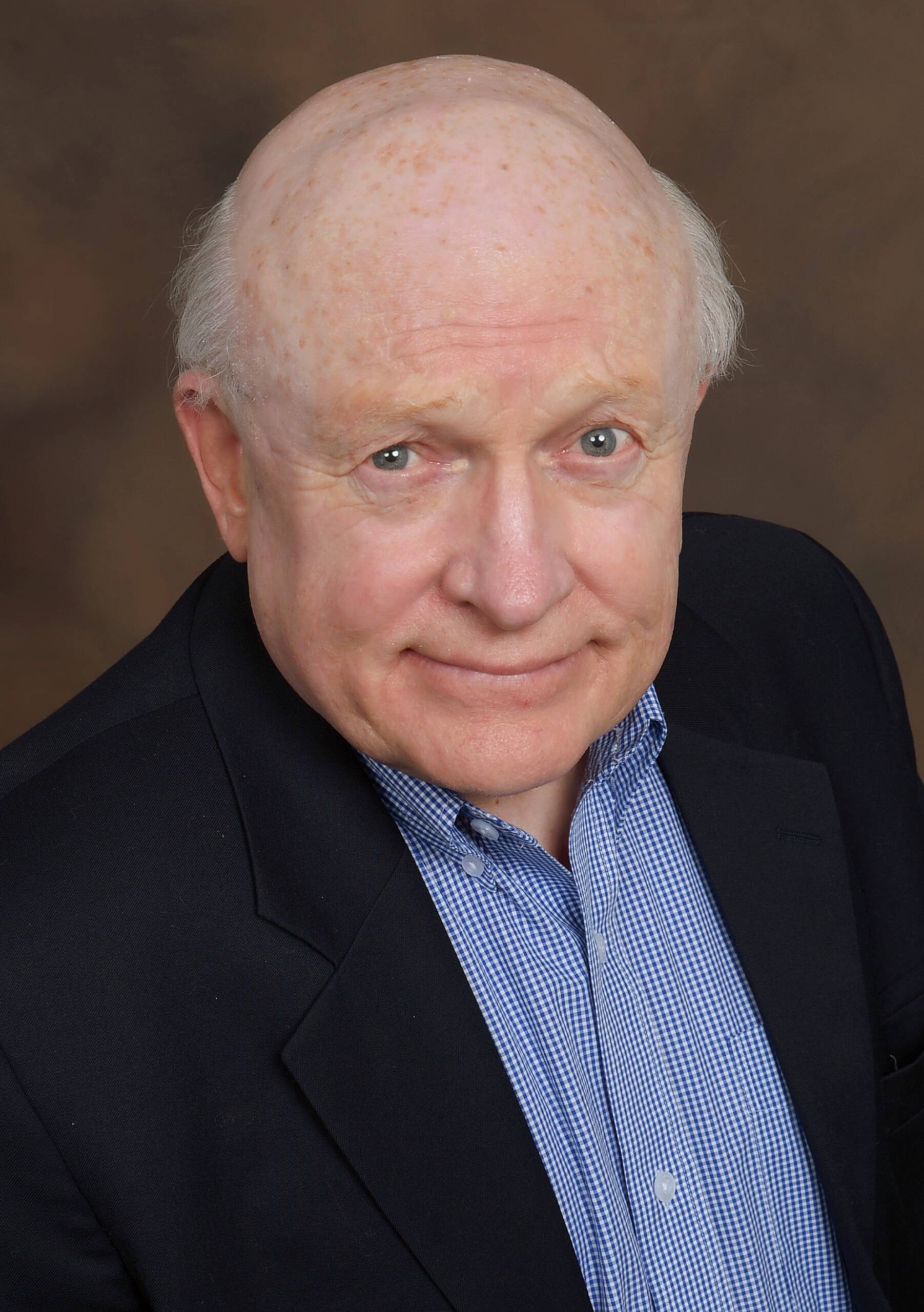 An older man with balding hair and a friendly expression, wearing a dark suit jacket and a light blue striped shirt, poses for a portrait against a brown background.