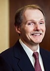 A man with light skin, short brown hair, and a mustache is smiling. He is wearing a dark suit jacket, a white shirt, and a red tie. The background is softly blurred with brown tones.