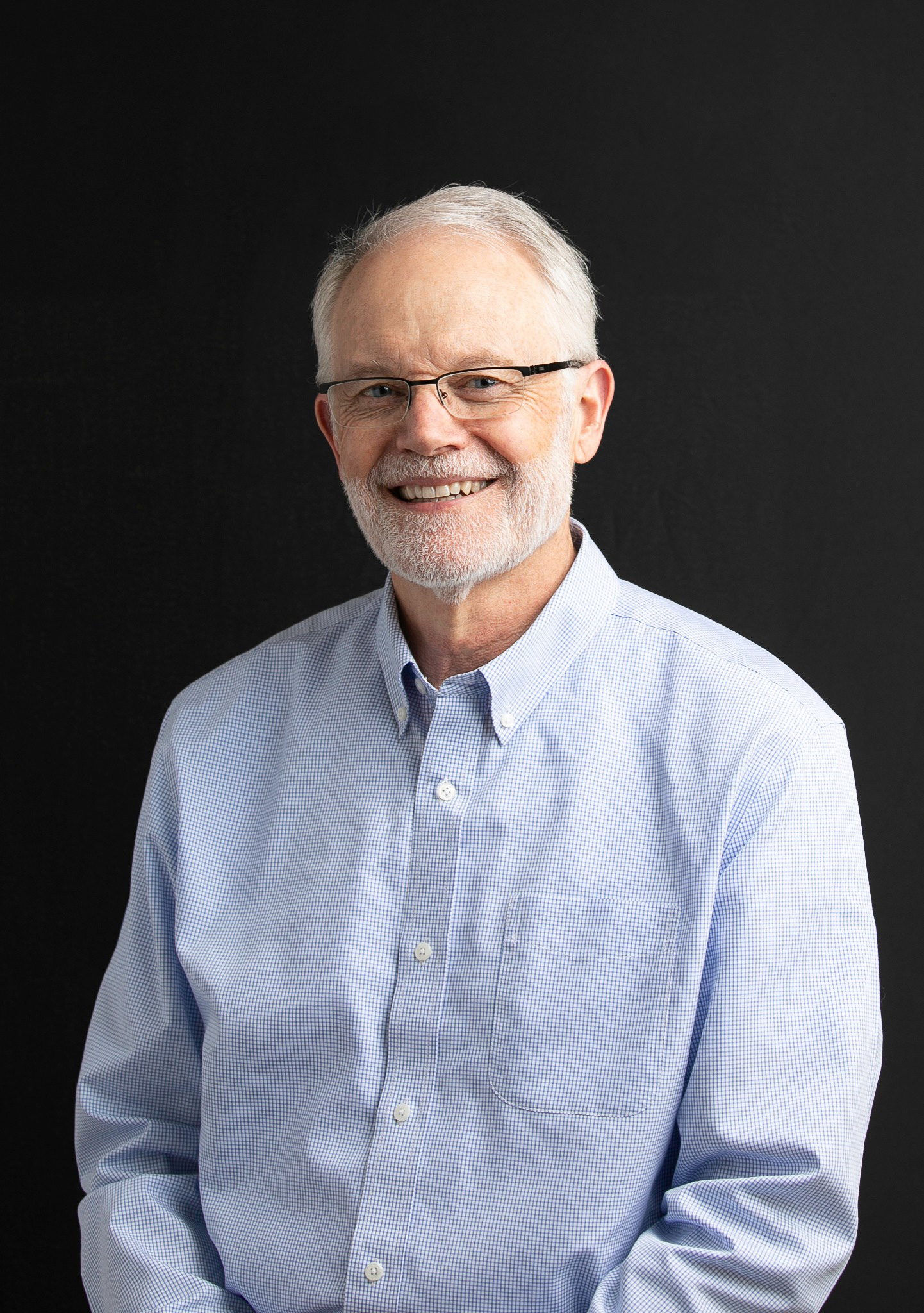 An older man with short white hair, glasses, and a trimmed beard smiles at the camera. He is wearing a light blue button-up shirt and is posed against a plain black background.