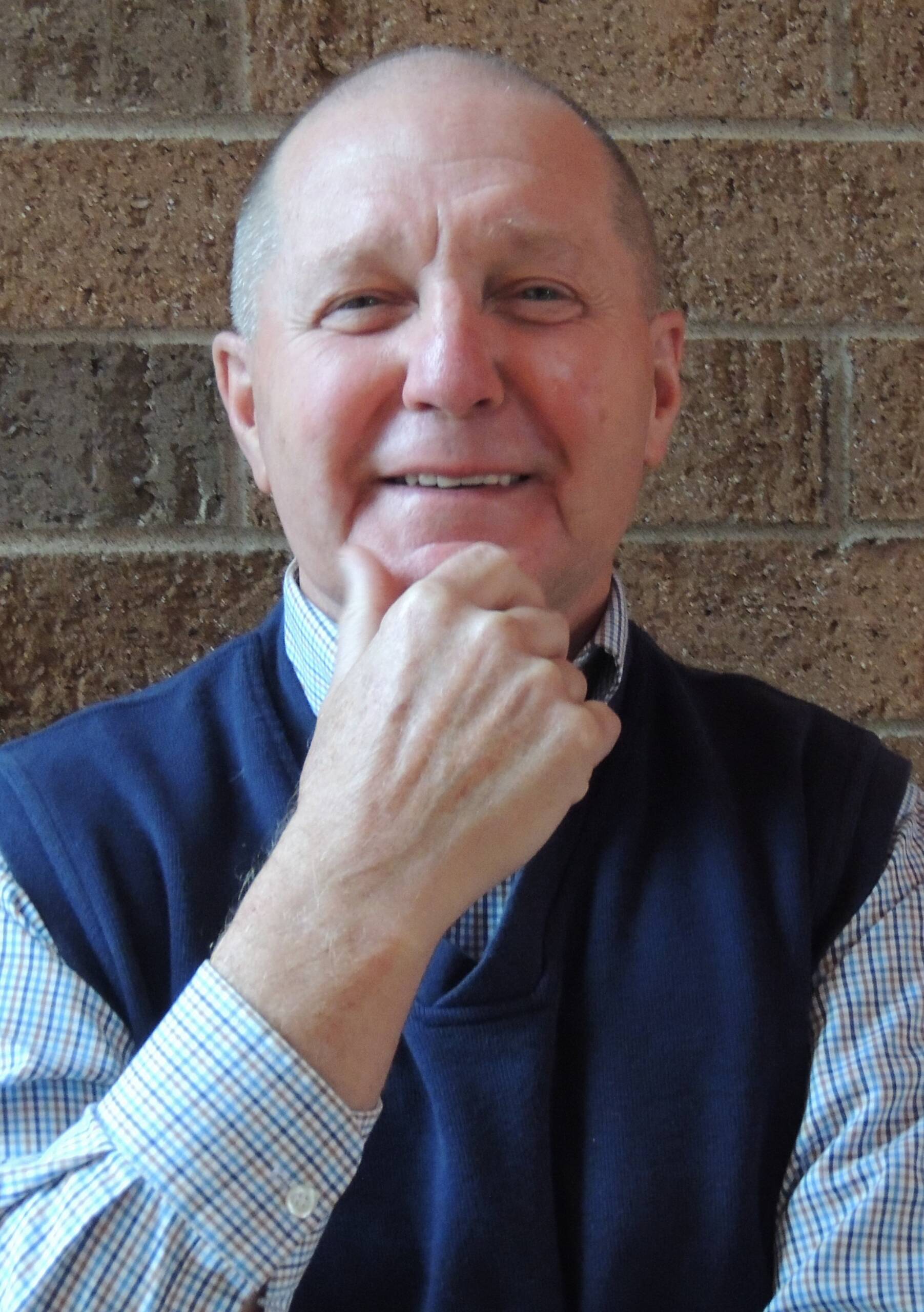 A person with short hair, wearing a blue sweater vest over a checked shirt, smiles while resting their chin on their hand, sitting in front of a brick wall.