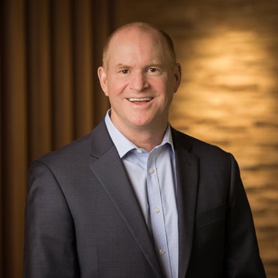 A smiling man in a business suit and light blue shirt stands in front of a softly lit background with wooden panels and a textured wall.