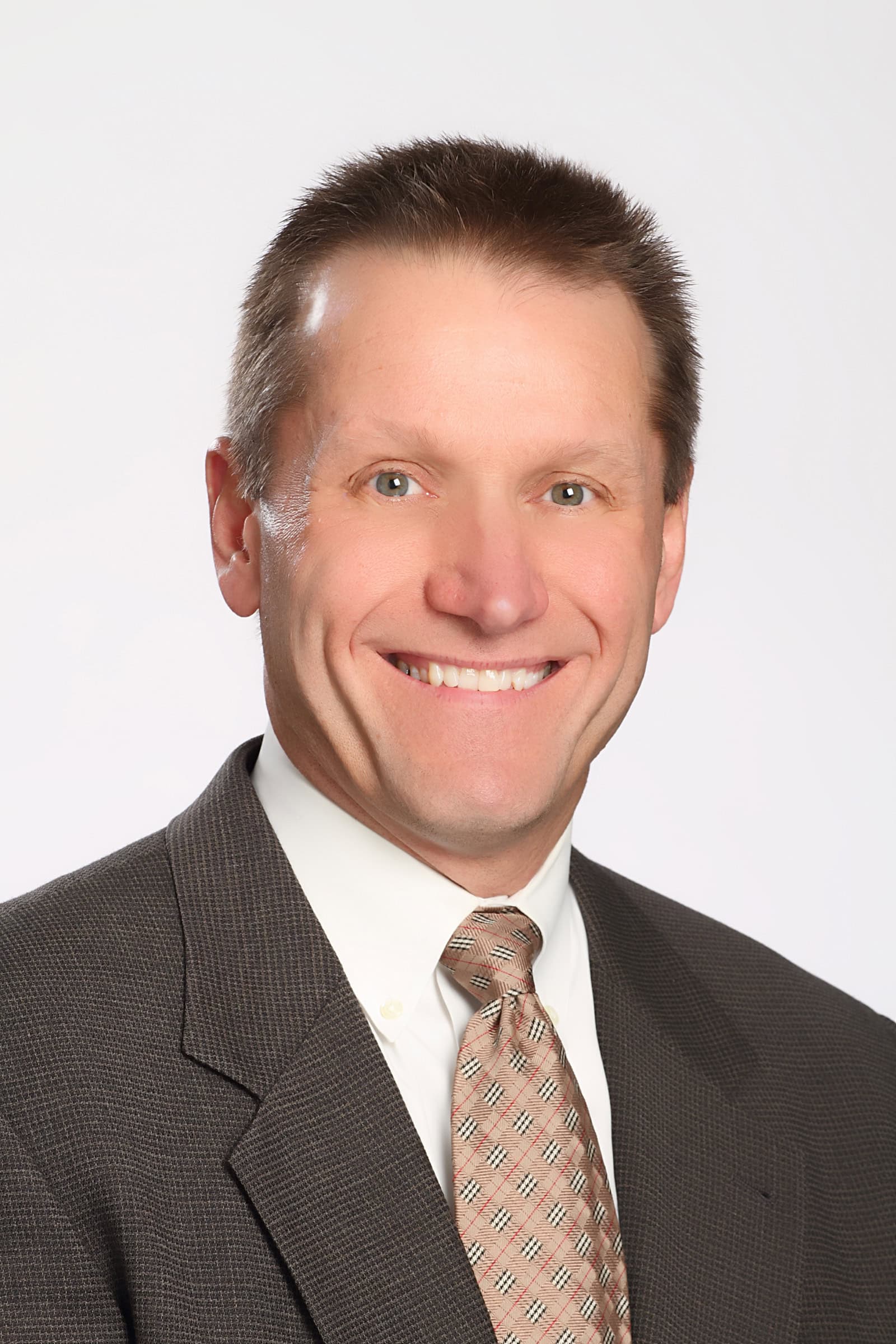 A man in a dark suit, white shirt, and patterned tie smiles at the camera against a plain light background. He has short, light brown hair and a fair complexion.