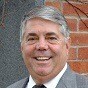 A smiling middle-aged man with gray hair wearing a gray suit and tie stands in front of a brick wall.