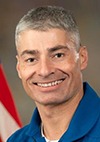 A man with short gray hair, wearing a blue collared shirt, is smiling at the camera. Part of an American flag is visible in the background.
