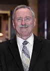 A smiling older man with gray hair and a mustache is wearing a dark suit, white shirt, and patterned tie, standing indoors in a well-lit setting.