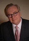 A man with light hair and glasses, wearing a dark pinstripe suit, white shirt, and pink tie, poses in front of a plain, neutral background. He is looking at the camera with a slight smile.