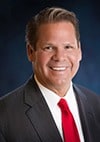 A man in a dark suit, white shirt, and red tie smiles at the camera against a dark blue background.