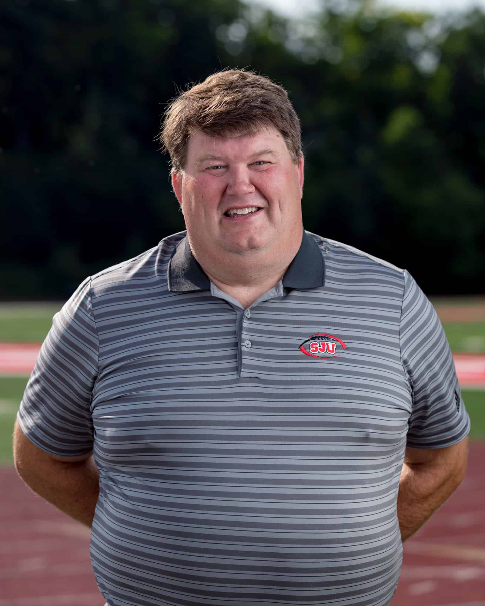 A man with short brown hair wearing a gray striped polo shirt with an "SJU" logo stands outdoors on a track field, smiling at the camera with his hands behind his back. Trees are visible in the background.