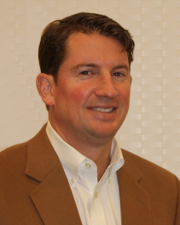 A man with short brown hair, wearing a tan blazer over a white collared shirt, is smiling in front of a light-colored, textured background.