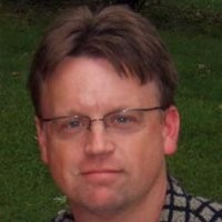 A man with short brown hair wearing glasses and a patterned shirt looks at the camera with a neutral expression. Green grass and blurred foliage are visible in the background.