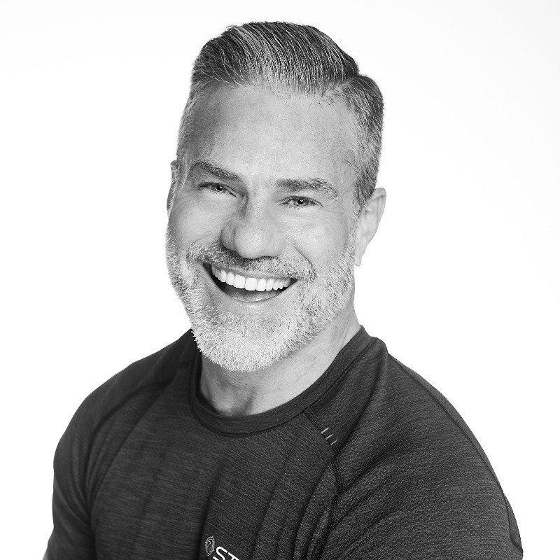 Smiling man with short, neatly styled gray hair and a beard, wearing a dark athletic shirt, posing against a plain white background.