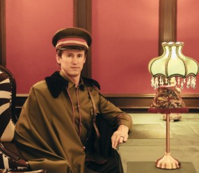 A man in a green military-style uniform and cap sits at a desk with a vintage green lampshade, set against a red and brown warmly lit background.