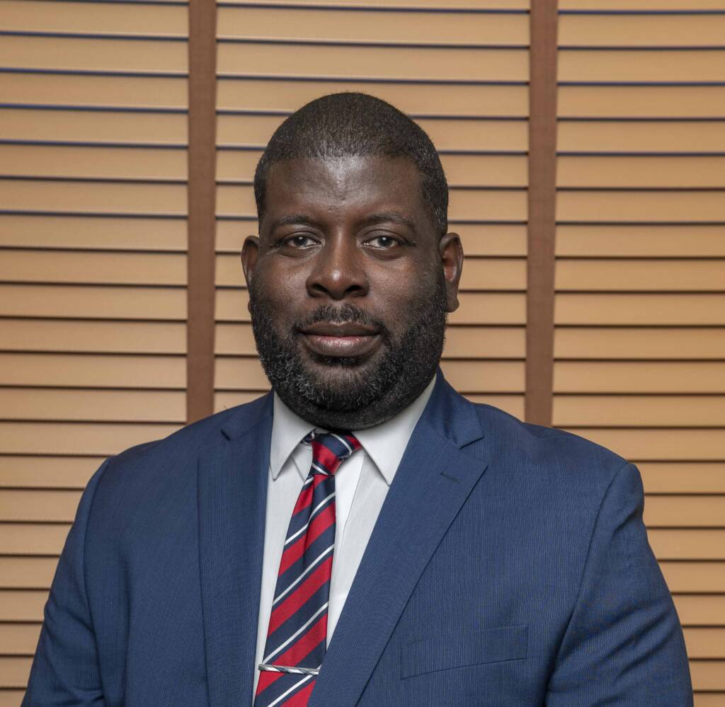 A man in a blue suit, white shirt, and red striped tie stands in front of wooden blinds, looking at the camera with a neutral expression.