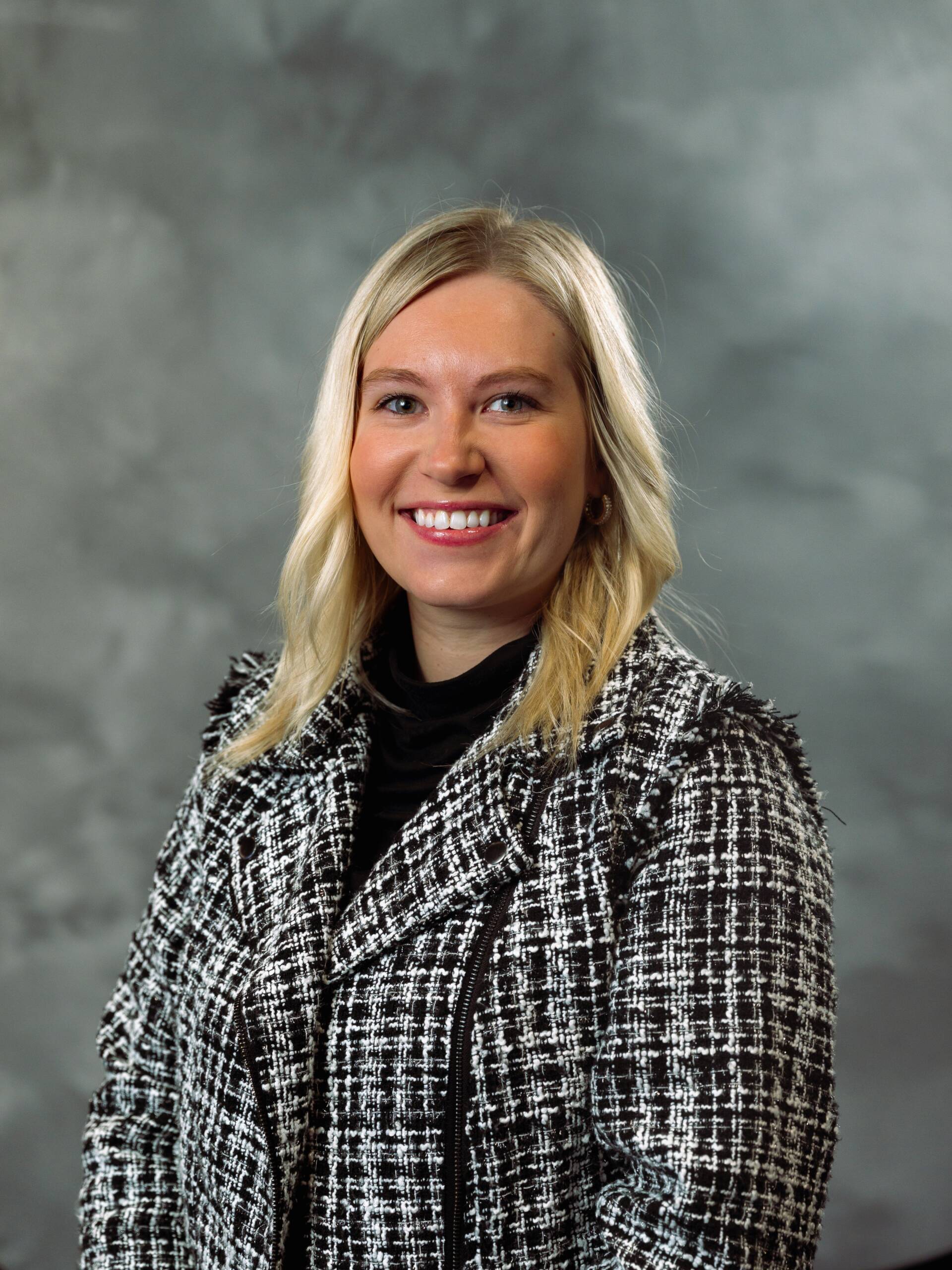 A woman with blonde hair wearing a black and white patterned jacket and a black top smiles at the camera against a gray, cloudy background.