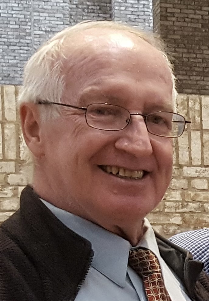 An older man with short white hair, glasses, and a patterned tie smiles at the camera. He is indoors, with a stone wall and brick background.