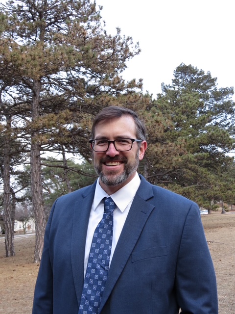 A man with glasses and a beard, wearing a blue suit and patterned tie, stands outside in a park with tall pine trees and brown grass in the background.