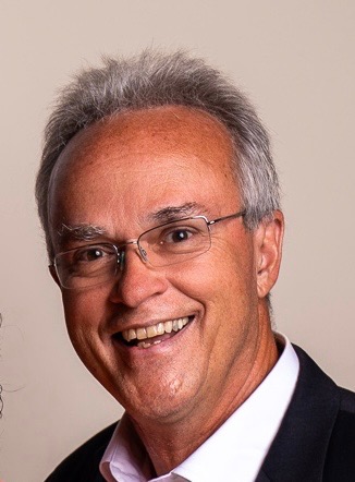 Smiling older man with short gray hair and glasses, wearing a dark blazer and a white shirt, posing in front of a plain beige background.