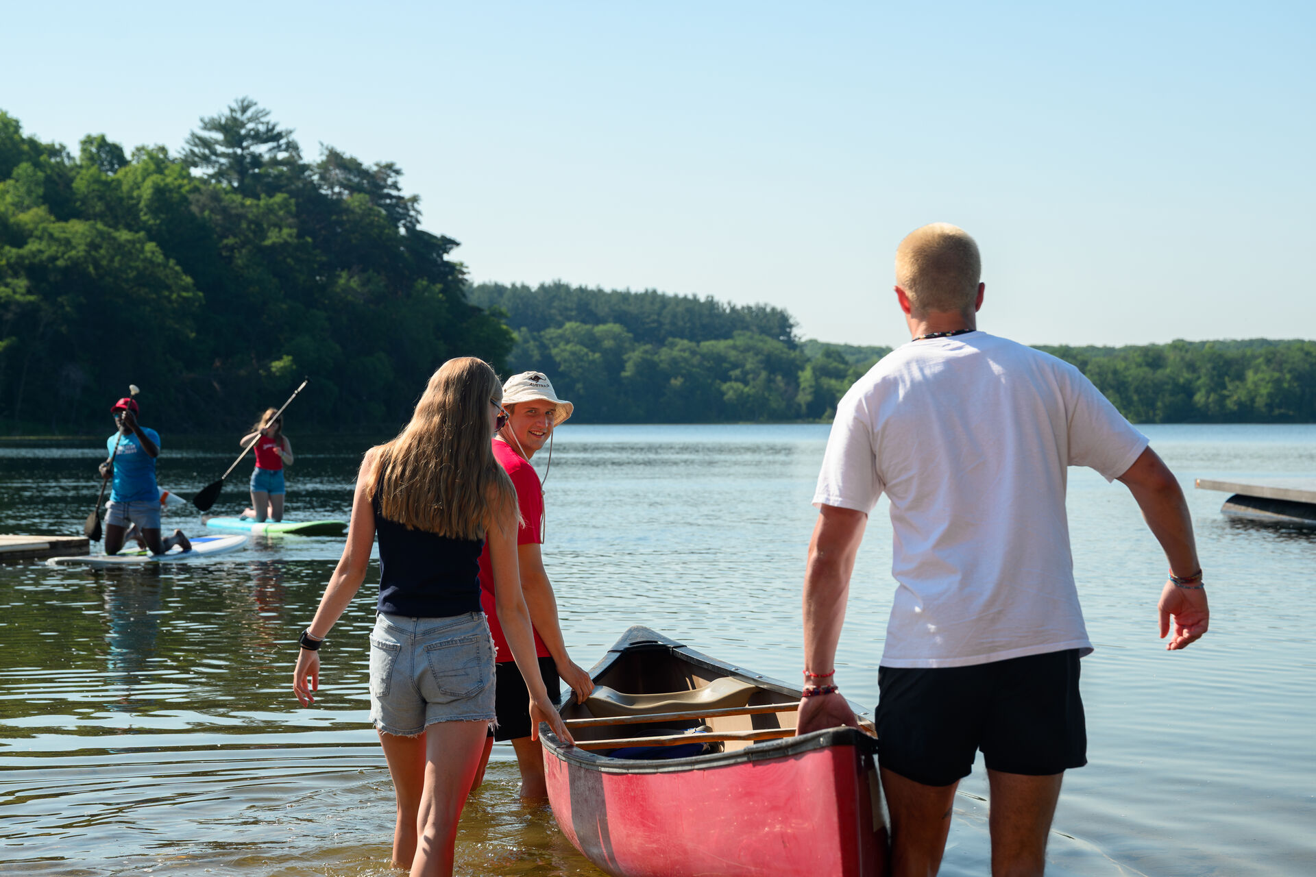 A group of young people stand at the edge of a lake, preparing to launch a red canoe. Two people paddle on stand-up paddleboards in the background, surrounded by trees under a clear sky.