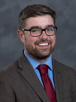 A man with short brown hair and a beard, wearing glasses, a brown suit jacket, blue shirt, and red tie, smiles in front of a gray, mottled studio background.