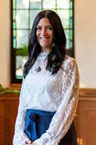 A woman with long dark hair wearing a white lace blouse and blue skirt stands indoors, smiling in front of a stained glass window. There’s a potted plant to her left and wood paneling in the background.