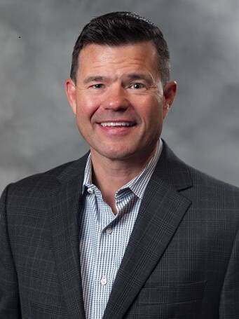 A man with short dark hair, wearing a gray checked suit jacket over a light blue patterned shirt, smiles at the camera in front of a gray, cloudy background.