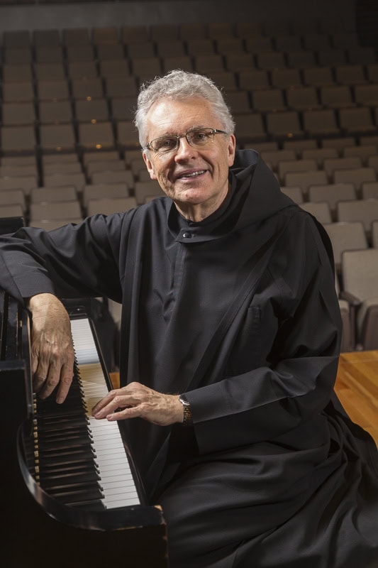 An older man wearing black religious robes sits at a grand piano, smiling at the camera. Empty, tiered seats are visible in the background, suggesting an auditorium or concert hall.
