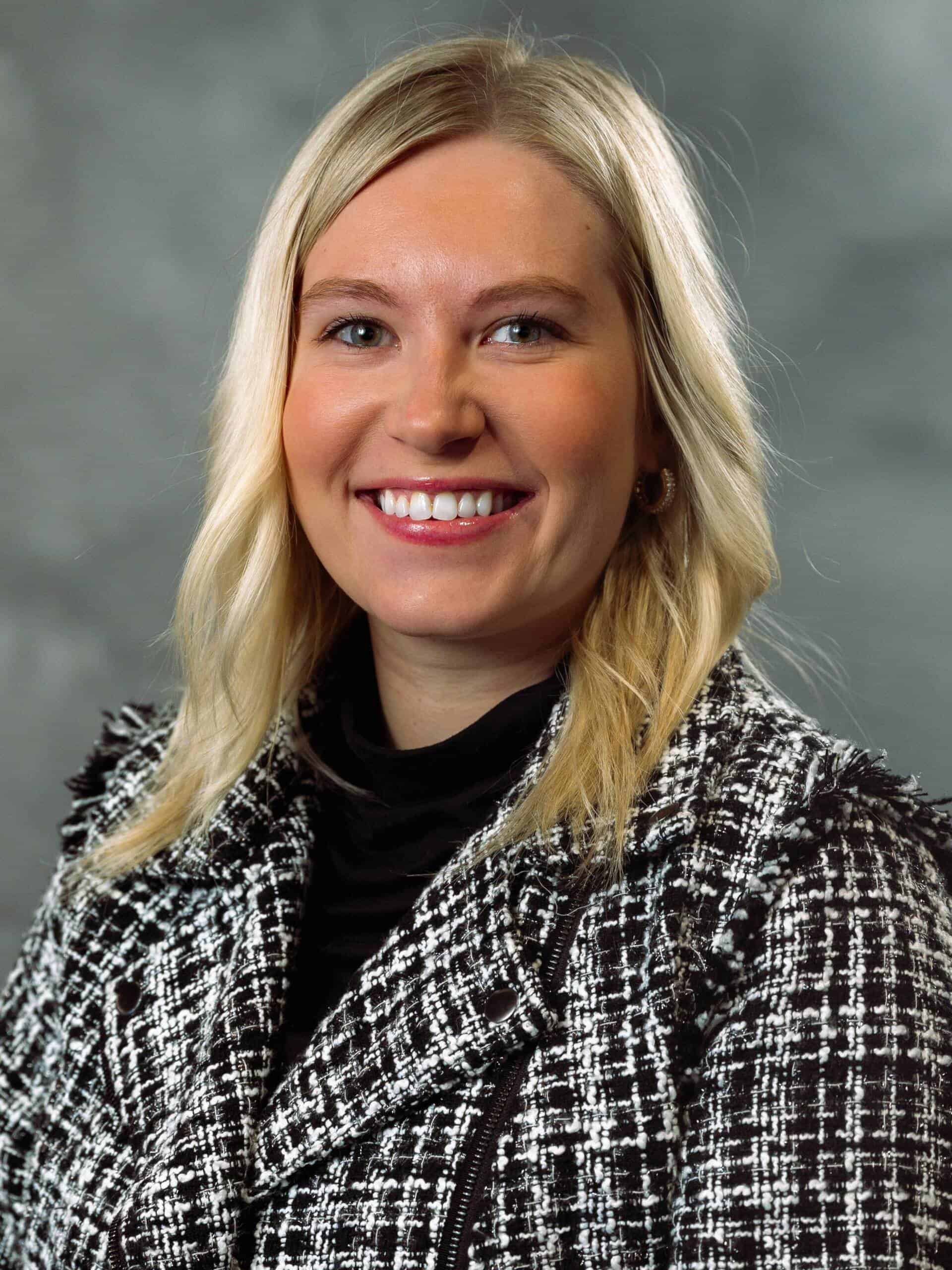 A woman with blonde hair wearing a black and white patterned jacket and a black top smiles at the camera against a gray, cloudy background.