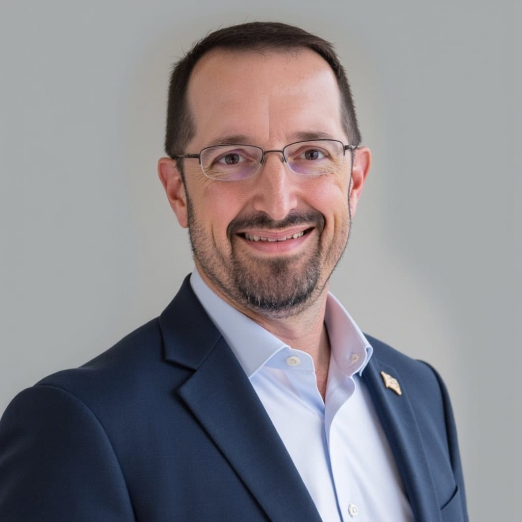 A man with short dark hair, a beard, and glasses is smiling. He is wearing a navy blue suit jacket, a white shirt, and an American flag pin, standing in front of a plain light gray background.