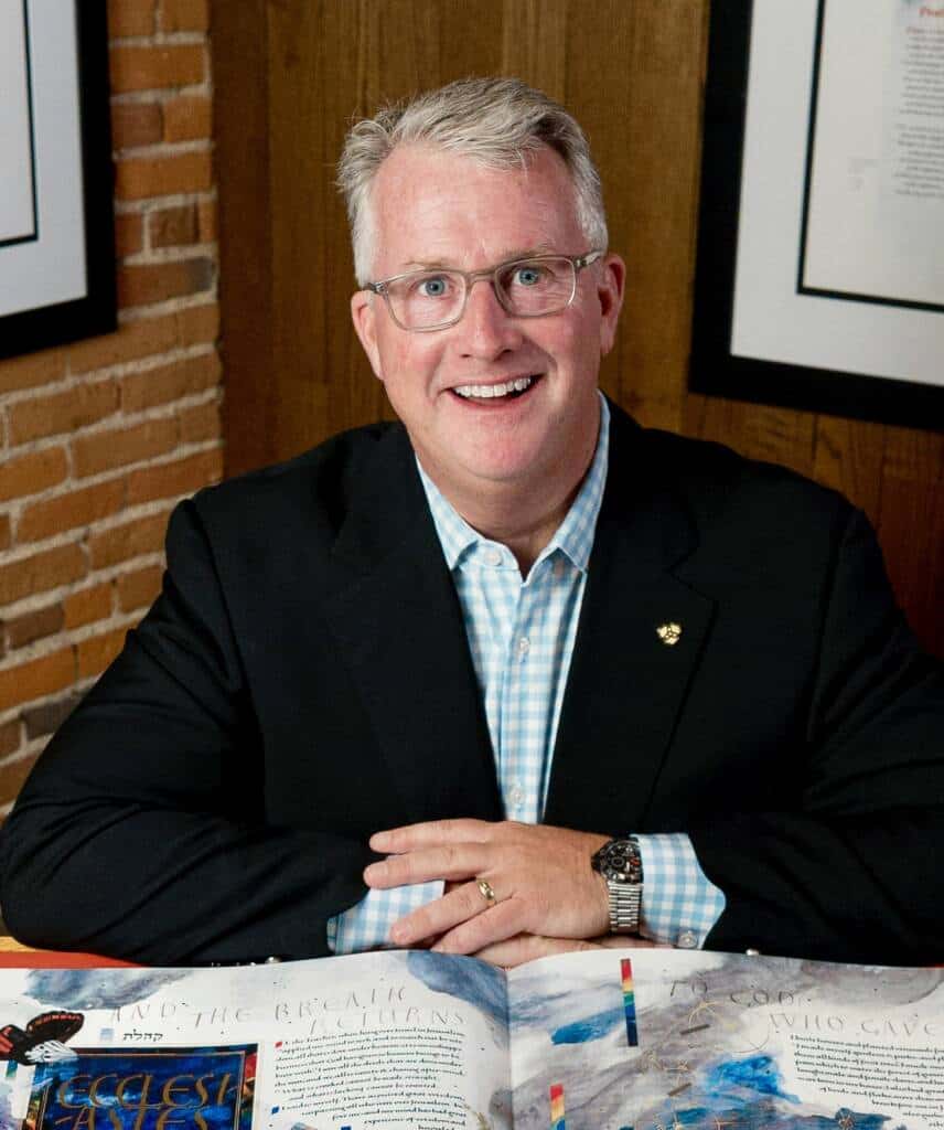 A smiling man in a suit and checkered shirt sits at a table with an open colorful book in front of him. He is in a room with wood paneling, brick walls, and framed pictures on the walls behind him.