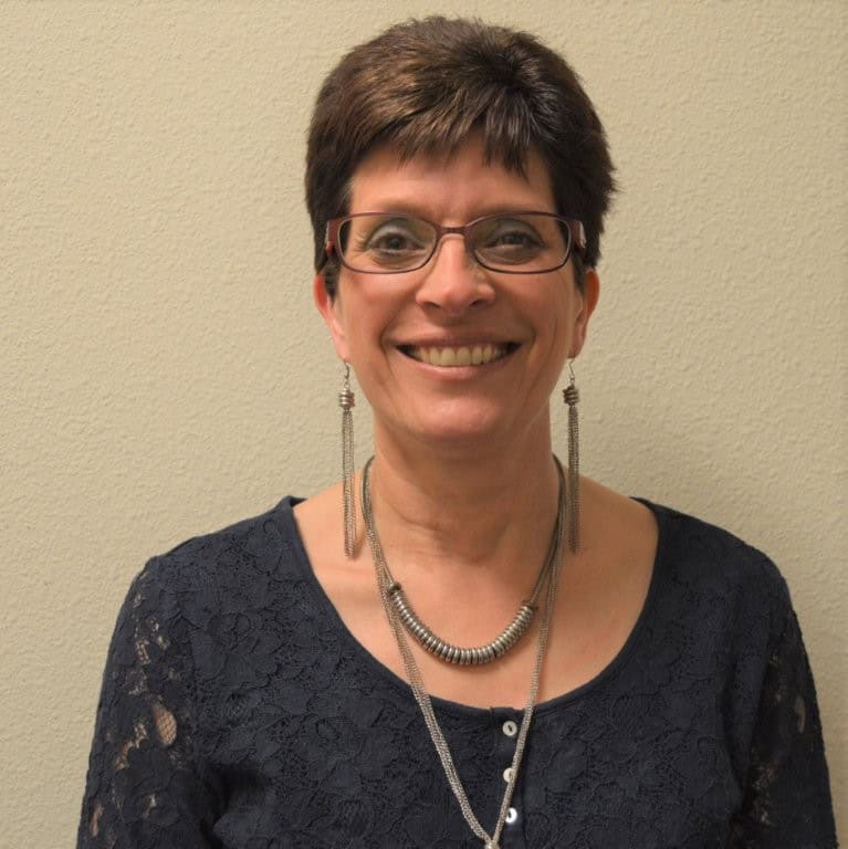 A woman with short brown hair and glasses smiles at the camera. She is wearing a dark lace top, long silver earrings, and layered silver necklaces, standing against a plain beige wall.