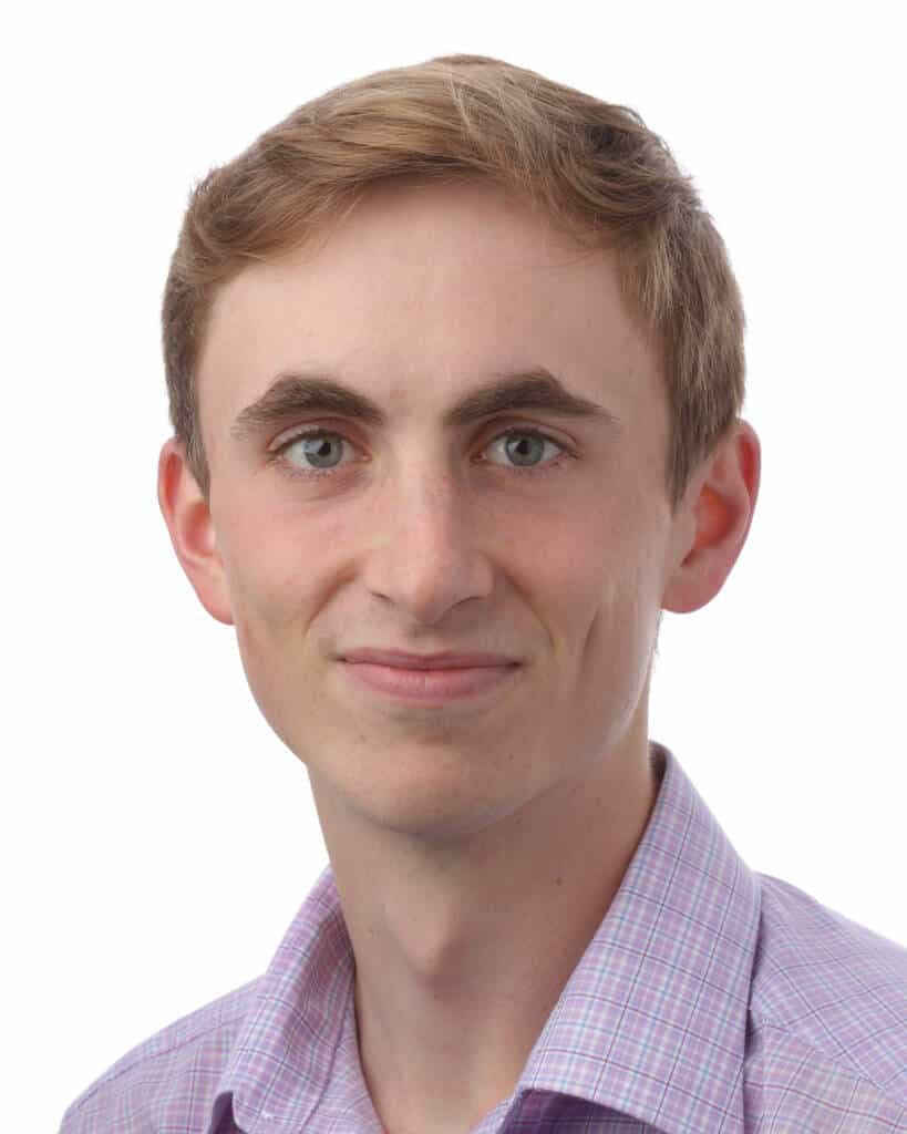 Young person with short light brown hair and blue eyes, wearing a light purple checkered shirt, smiling slightly while looking at the camera, against a plain white background.