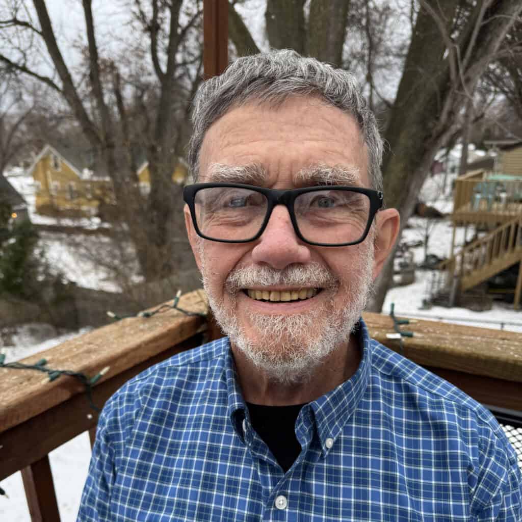 An older man with gray hair, a beard, and glasses smiles at the camera while standing on a wooden deck. In the background, there are leafless trees and snow-covered ground and houses.