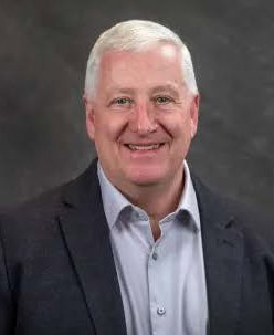 A middle-aged man with short white hair, wearing a light gray dress shirt and a dark blazer, smiles at the camera against a plain dark background.