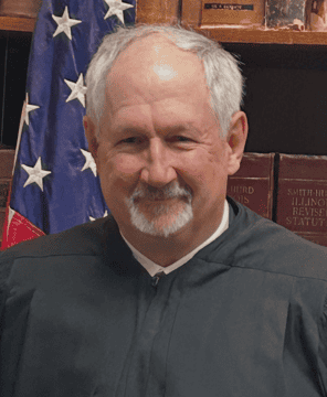 A smiling older man with short gray hair and a mustache, wearing a black judge's robe, stands in front of a U.S. flag and law books.