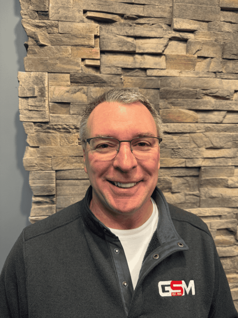 A smiling man with short gray hair and glasses is wearing a dark pullover with a “GSM” logo, standing in front of a wall with a textured stone pattern.