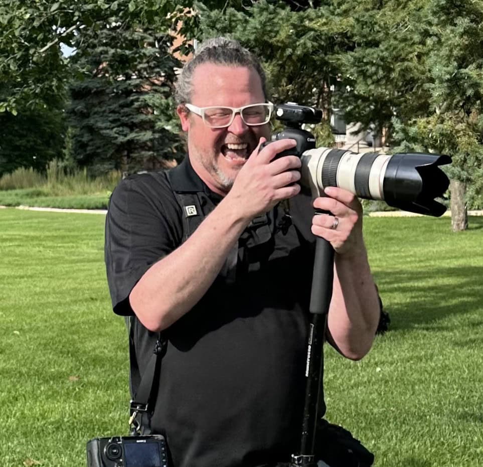 A man with glasses and a beard smiles while looking at a camera with a large telephoto lens, standing on a grassy area with trees in the background.
