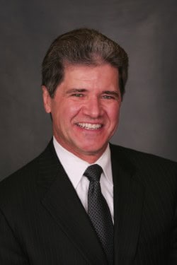 A man in a dark pinstripe suit, white shirt, and patterned tie is smiling against a plain dark background.