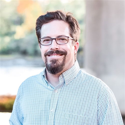 A man with short dark hair, glasses, and a beard is smiling and wearing a checkered button-down shirt. The background is outdoors with a soft focus on greenery and a body of water.