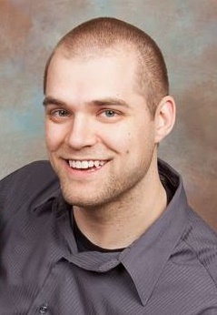 A young man with a buzz cut smiles at the camera. He is wearing a dark gray button-up shirt, and the background is a gradient of muted colors, primarily shades of brown and blue.