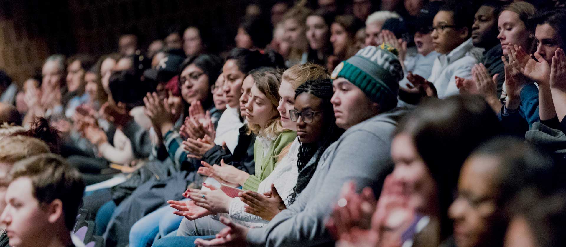 A diverse group of people sitting closely together in an auditorium, attentively watching something off-camera and clapping. They appear to be of various ages and are dressed in casual clothing. The atmosphere is serious and focused.