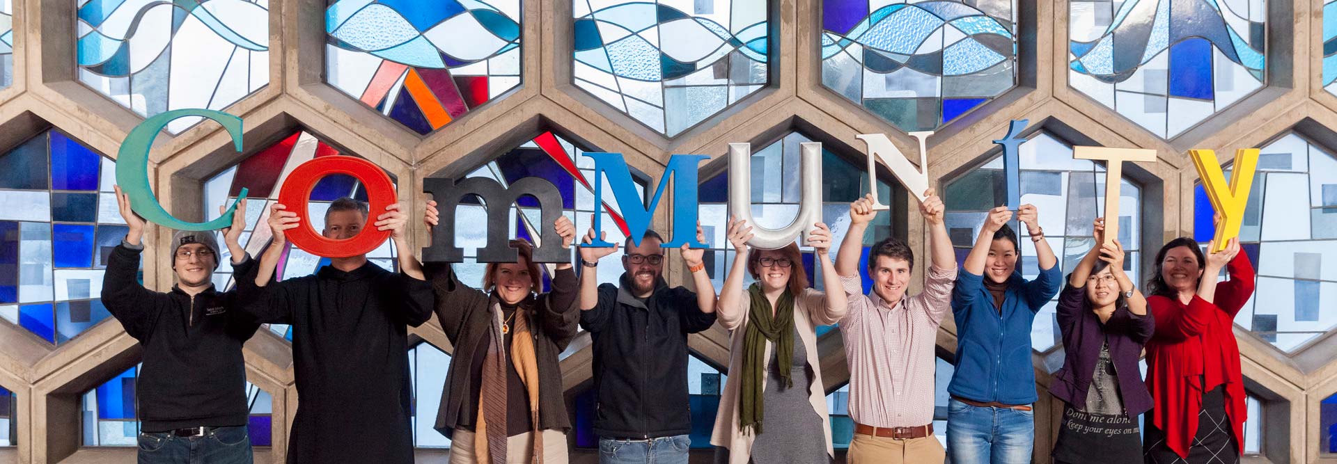 A diverse group of eight people stand in front of a colorful stained glass window, each holding up a large, brightly colored letter that together spell "COMMUNITY." They appear cheerful and unified, celebrating togetherness.